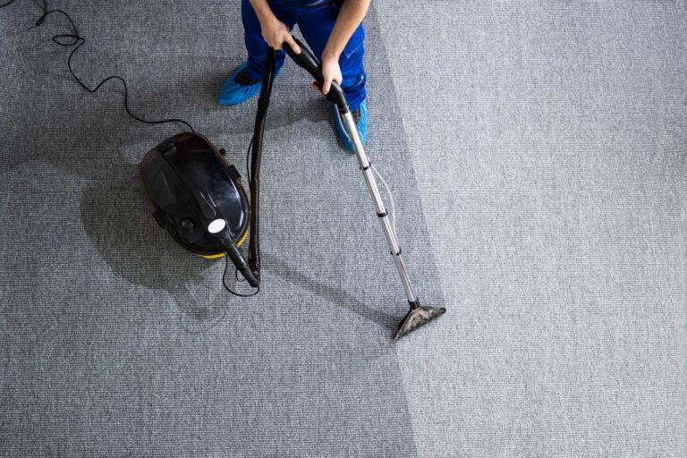 A person wearing blue overalls is using a vacuum cleaner on a carpet, focusing on cleaning the surface.