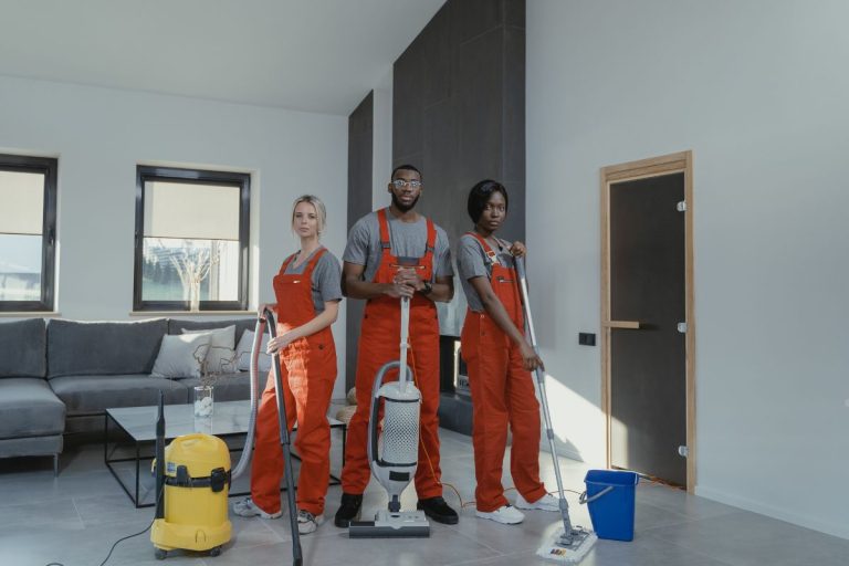 Three individuals in overalls are actively cleaning a room, using various cleaning tools and equipment.